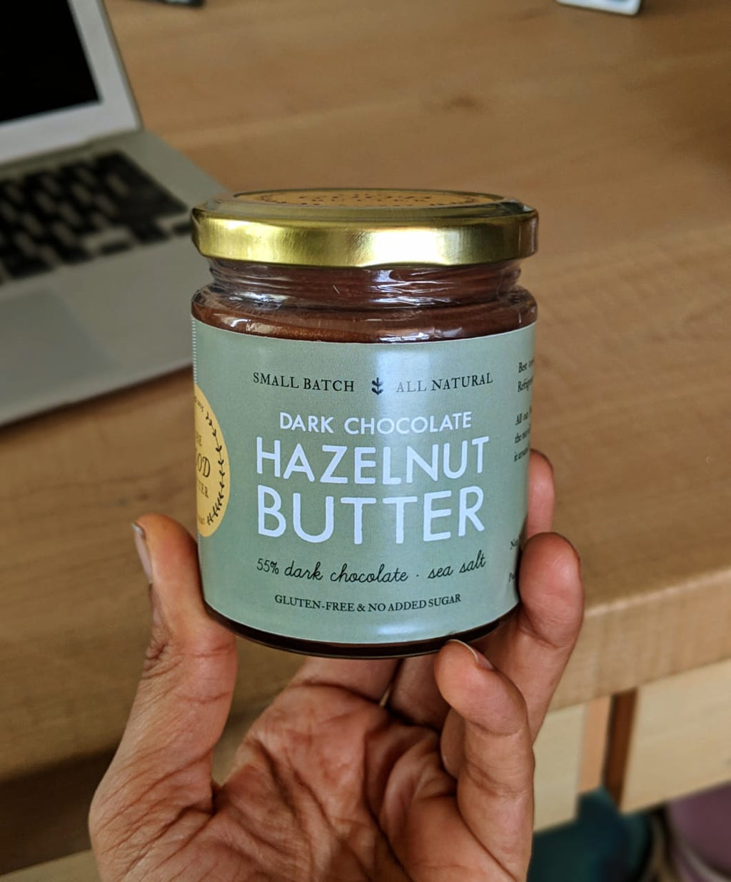 A jar of Hazelnut Dark Chocolate Butter being held by a person, with the label displaying the product name and details, placed on a wooden surface.